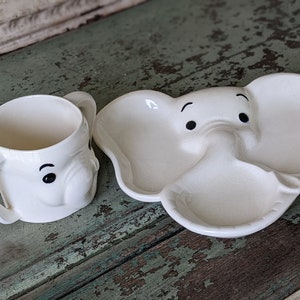 May include: A white ceramic elephant-shaped plate with a divided section for food. The plate has a cute cartoon elephant face with black eyes and a trunk. A matching white ceramic elephant-shaped mug with black eyes is also included.