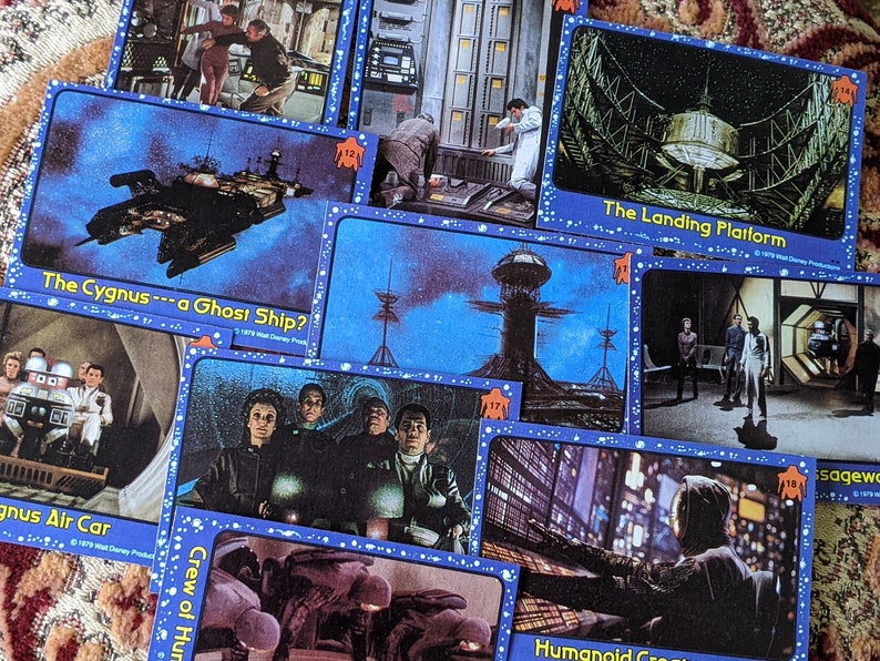 May include: A collection of trading cards featuring images from the 1979 film 'The Black Hole'. The cards feature images of the spaceship, the landing platform, the crew, and the air car. The cards are blue with white stars and have the title of the card printed in white.