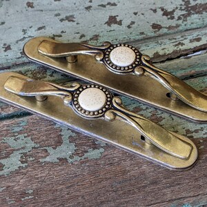 May include: Two antique brass drawer pulls with ornate details and white ceramic centers.