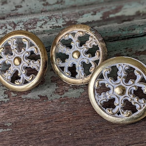 May include: Three round, decorative, gold-colored metal knobs with a white, ornate, floral design. The knobs have a raised, circular center.