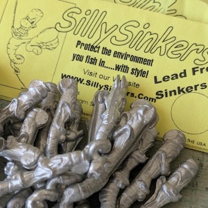 May include: A pile of silver-colored metal fishing sinkers in front of a yellow card with the text SillySinkers, Protect the environment you fish in...with style!, Lead Free Sinkers, and a cartoon illustration of a worm.