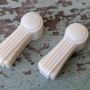 May include: Two off-white, vintage-style window cranks. Each crank features a circular head with concentric rings and a ribbed handle. The cranks are set against a distressed, teal-colored wooden surface.