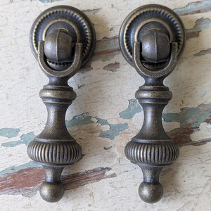 May include: Two matching antique brass-toned metal drop pulls featuring a circular backplate with a ridged edge, a hinged bail, and a vertical stem with a ribbed bulbous base ending in a small sphere, designed for furniture drawer or cabinet hardware.