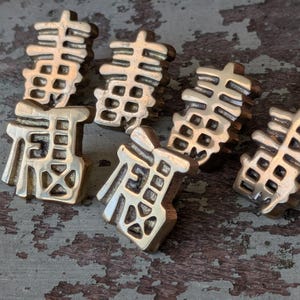 May include: Six bronze-colored charms, each featuring a stylized Chinese character. The charms are arranged on a distressed, textured surface with a mix of brown and gray tones. The characters are detailed and three-dimensional.