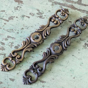 May include: Two ornate, antique-style drawer pulls. One is a warm brass color, the other is a darker bronze. Each has a scrolling, floral design and two screw holes for mounting. These decorative pulls are ideal for furniture restoration projects.