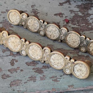 May include: Two antique brass cabinet pulls with a series of circular, textured inserts. The pulls have a warm, golden hue and are arranged on a weathered, teal-colored wooden surface. Each pull features multiple circular designs.