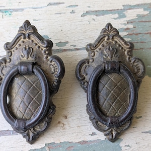 May include: Two matching drawer pulls featuring an ornate, antiqued brass-toned metal backplate with a diamond-patterned center and a dark, circular drop handle. The hardware displays intricate scrollwork details and a weathered, aged finish.