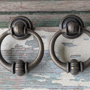 May include: Two antique bronze-colored ring pulls for furniture. Each pull features a circular ring attached to a decorative backplate. The ring has a central, ribbed design with rope-like detailing. The backplates have a circular, ridged design.
