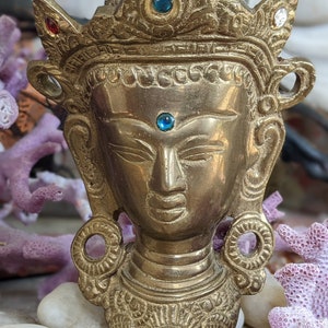 May include: A gold-colored brass statue of a deity with a crown and blue and red gemstones. The statue has a serene expression and is decorated with intricate details.