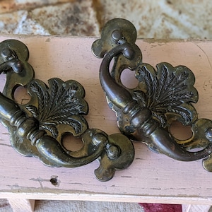 May include: Two antique brass cabinet pulls with ornate floral designs. The pulls have a curved handle and a decorative floral design with a central leaf motif.