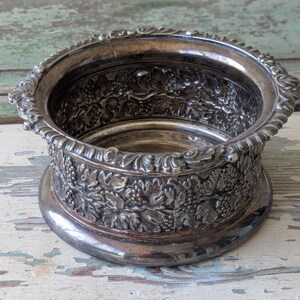 May include: Silver-toned wine coaster with an ornate design. The coaster features a detailed relief of grape vines and leaves around the sides. The rim has a decorative, scalloped edge. The coaster sits on a rounded base.