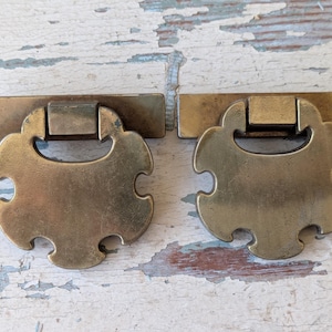 May include: Two antique brass drawer pulls. Each pull has a rectangular backplate and a decorative, scalloped handle. The brass has a warm, aged patina, suggesting vintage or antique hardware. The pulls are designed for furniture restoration or decorative use.