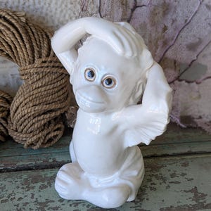 May include: A glossy white ceramic figurine of a seated orangutan with its right hand raised to its forehead and its left hand resting near its ear. The primate has detailed facial features with dark, expressive eyes and a smooth, reflective finish.