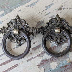 May include: Two matching metal drawer pulls featuring a dark bronze finish. Each pull consists of a decorative, lattice-patterned backplate with ornate scrollwork edges and a circular ring handle attached to a central mounting point.