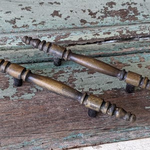 May include: Two antique brass cabinet door pulls with a decorative design. The pulls are on a weathered wooden surface.