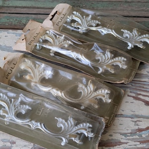 May include: Four vintage cabinet pulls with an ornate, white floral design, still in their original packaging. The pulls are made of metal and have a gold-colored accent. The packaging is a faded tan color with the word "PULL" printed on the top.