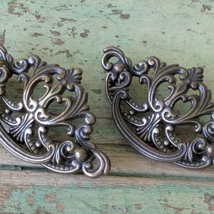 May include: Two antique bronze-colored corner cabinet decorative hardware pieces. Each piece has an ornate, symmetrical design with swirling patterns and small ball accents. The hardware is designed to be mounted on a corner.