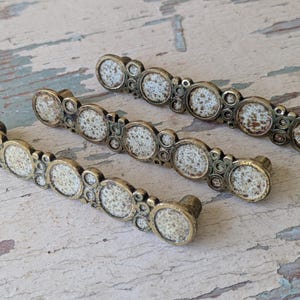 May include: Three antique-style cabinet pulls with a distressed finish. Each pull features a series of circular, off-white speckled inserts set within a decorative, aged brass-toned metal frame. The pulls are designed for easy installation.