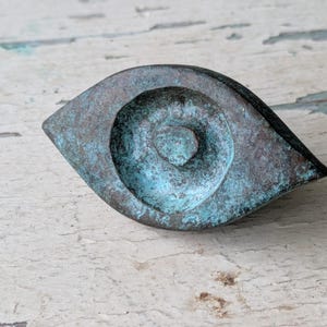 May include: An eye-shaped object with a weathered, oxidized finish in shades of dark brown and teal green. The center features a raised, circular pupil detail, creating a textured, metallic appearance with a distinct almond-shaped outline.