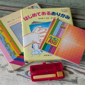May include: A collection of Japanese origami books with colorful covers and text. A red case with a yellow object is in the foreground. The books are stacked on a weathered wooden surface.