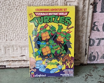 May include: A box of Teenage Mutant Ninja Turtles Colorforms, featuring the four turtles in action poses. The box is yellow with a black and green image of the turtles. The text on the box reads "Colorforms Adventure Set" and "Teenage Mutant Ninja Turtles".