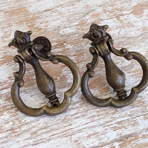 May include: Two antique brass-toned metal drawer pulls featuring a drop-style handle with a decorative crown-shaped top mount and a curved, ornate loop design. These hardware pieces have a weathered, textured finish and a central vertical post.