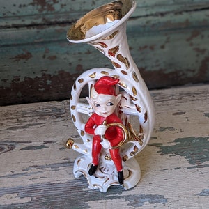 May include: A white ceramic figurine of a Christmas elf playing a golden trumpet. The elf is wearing a red hat and suit with white trim. The trumpet is decorated with gold accents.