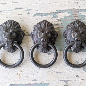 May include: Three dark bronze-toned metal drawer pulls, each featuring a detailed lion head design with a circular ring handle suspended from the lion's mouth. These hardware pieces have a weathered, aged finish and a classic, ornate appearance.