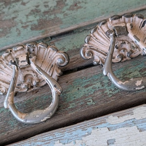 May include: Two antique-style drawer pulls with a distressed finish. Each pull features a decorative, fan-shaped backplate in off-white and a curved, silver-toned handle. The pulls are set against a weathered, multi-colored wooden surface.