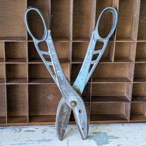May include: A pair of vintage metal shears with a silver finish. The shears are open and resting on a wooden surface with multiple compartments.