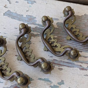May include: Four antique brass drawer pulls with ornate floral designs. Each pull features a curved handle and decorative backplate. The pulls are a warm brass color, showing signs of age and wear, and are set against a distressed, weathered wood surface.