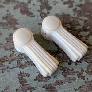 May include: Two vintage ivory-colored window crank handles. Each handle has a circular head with concentric rings and a ribbed, rectangular body. The handles are set against a distressed, weathered, teal-colored surface.