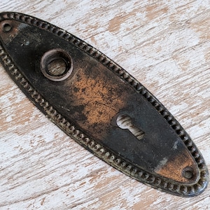 May include: An oval-shaped metal door plate with a dark, weathered patina and a beaded border. The plate features a central circular opening for a knob, a lower keyhole cutout, and two small screw holes at the top and bottom ends of the piece.
