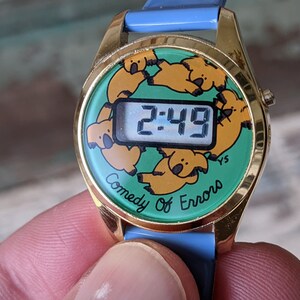 May include: A gold-tone digital watch with a green face featuring a repeating pattern of cartoon koalas. The watch face reads "2:49" and the text "Comedy of Errors" is printed below the digital display.
