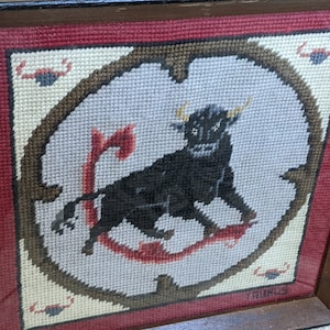 May include: A framed needlepoint embroidery depicting a black bull with red and brown accents. The embroidery is surrounded by a white border and a brown frame. The word "Taurus" is stitched in the bottom right corner.