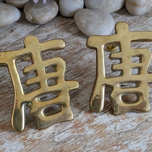 May include: A pair of gold-toned metal earrings shaped like the Chinese character Tang. Each earring features a polished, reflective surface with smooth, rounded edges and a post-style backing, displaying the intricate calligraphic symbol clearly.