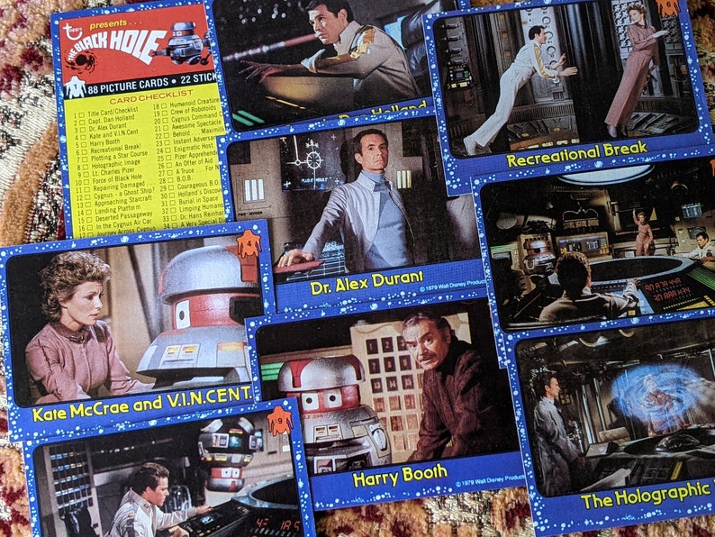 May include: A collection of trading cards featuring characters and scenes from the 1979 science fiction film "The Black Hole". The cards feature images of the characters, including Kate McCrae, V.I.N.CENT., Dr. Alex Durant, Harry Booth, and others. The cards also feature scenes from the film, such as the recreational break and the holographic projection.