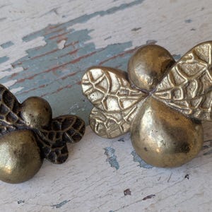 May include: Two antique brass bee-shaped cabinet knobs. The knobs have a detailed wing design and a rounded body. The brass has a warm, aged patina, and the knobs are set against a distressed, painted wooden surface.