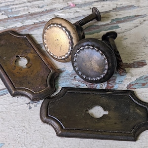 May include: Two antique brass cabinet knobs with a decorative edge and a keyhole plate. The knobs are round and have a slightly raised center. The plates are rectangular with rounded corners and a keyhole in the center.