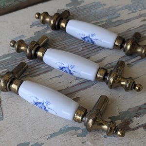 May include: Three drawer pulls featuring white ceramic centers with blue floral patterns and aged brass metal ends. Each handle has a cylindrical ceramic middle section decorated with delicate blue flowers and ornate brass hardware at both ends.