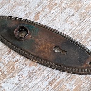 May include: An oval-shaped metal door plate featuring a dark, weathered patina with visible oxidation. The plate has a beaded border, a central circular opening for a knob, a keyhole below, and two small screw holes at the rounded ends.