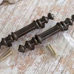 May include: Two antique brass cabinet door pulls with ornate details and a slightly aged finish. The pulls are shown with two screws in a plastic bag.
