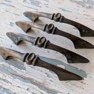 May include: Four antique bronze drawer pulls with a weathered patina. Each pull features a decorative center and a curved handle, ideal for adding a vintage touch to furniture. The pulls are arranged diagonally, showcasing their design.