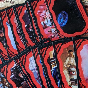 May include: A stack of trading cards with red and black borders. The cards feature images from the Star Wars movie franchise. The cards are in a variety of scenes and characters.