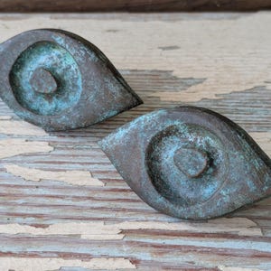 May include: Two antique bronze-colored, eye-shaped cabinet pulls with a teal patina. The pulls have a central circular design and are set against a weathered, light blue and brown wooden surface. These decorative hardware pieces add a vintage touch.