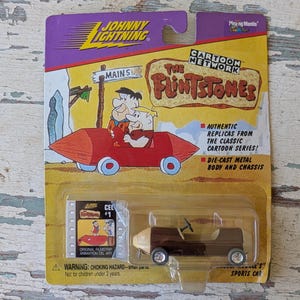 May include: A Johnny Lightning die-cast metal sports car from The Flintstones cartoon series, in its original packaging. The packaging features a cartoon image of Fred and Barney in their car, with the text "The Flintstones".