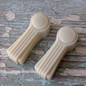 May include: Two off-white, vintage-style window cranks. Each crank features a circular head with concentric rings and a ribbed handle. The cranks are set against a weathered, distressed wooden surface with peeling paint.
