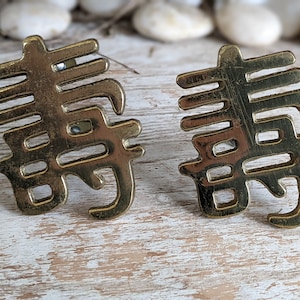 May include: A pair of brass-toned metal earrings shaped like the Chinese character for longevity, Shou. The earrings feature a polished, reflective surface with cut-out details forming the intricate strokes of the character in a symmetrical design.