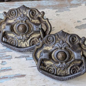 May include: Two antique bronze drawer pulls with ornate scrollwork and a central floral design. The pulls have a dark bronze finish with gold accents, and a small handle. These vintage hardware pieces are perfect for adding a touch of elegance to furniture.