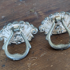 May include: Two antique drawer pulls with a distressed white and silver finish. Each pull features a decorative, shell-shaped backplate and a curved, teardrop-shaped handle. The hardware is likely made of metal.